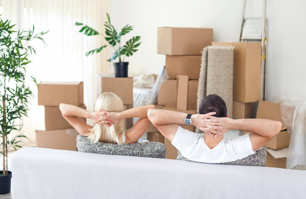 Un homme et une femme vus de dos assis dans un canapé et regardant des cartons de déménagement